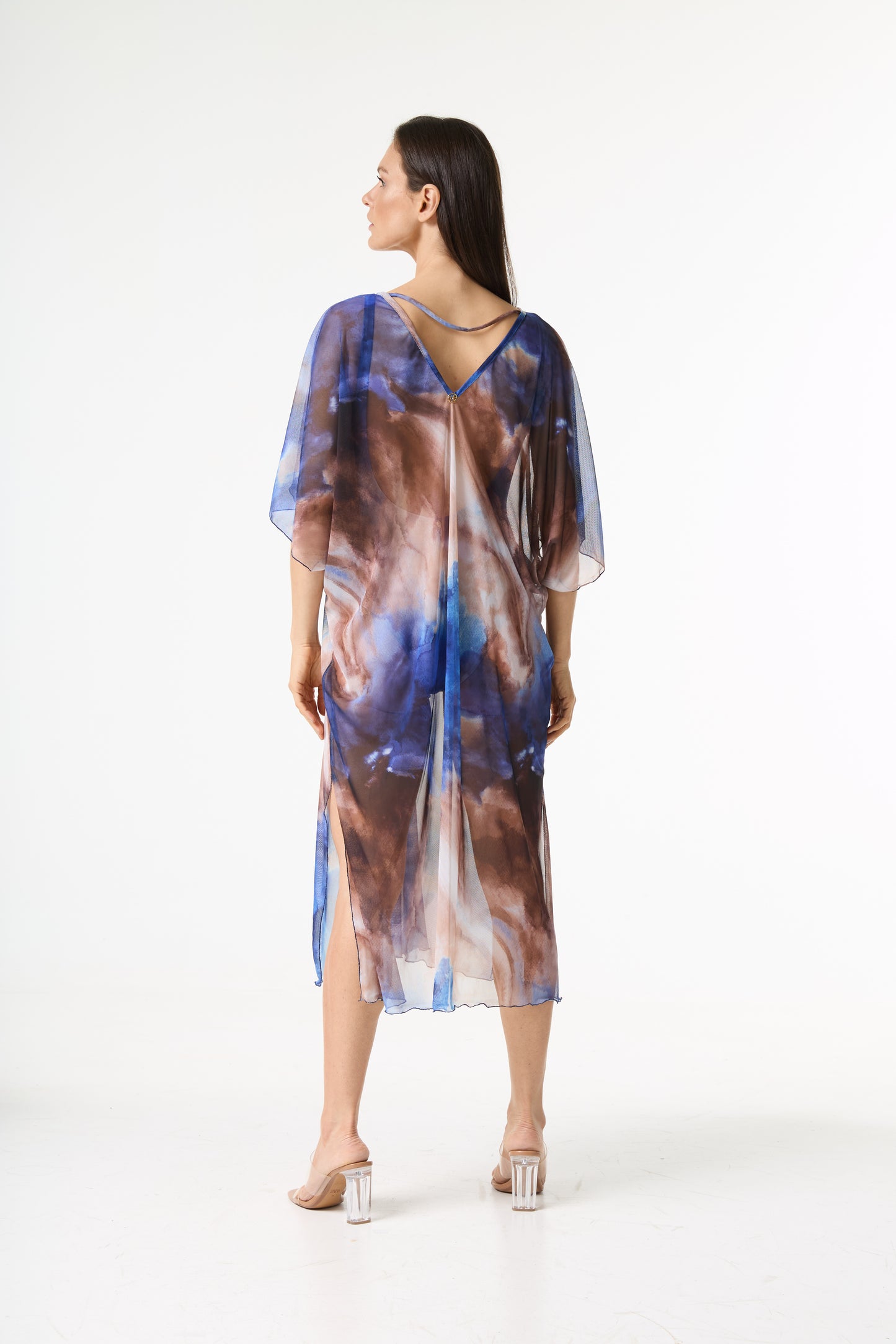 Saída de praia kaftan estampa tie dye. A modelo está de costas, olhando para o lado, exibindo o caimento fluido da peça transparente em tons de azul e marrom