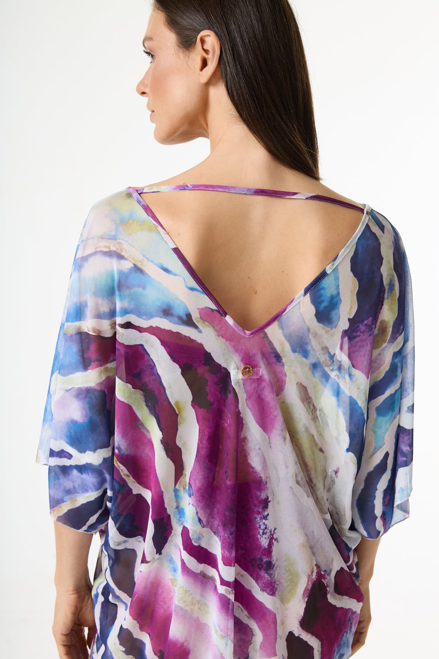 Blusa estampa abstrata colorida em tons de azul, roxo e magenta. A modelo posa de costas, exibindo o decote V profundo, a tira horizontal nas