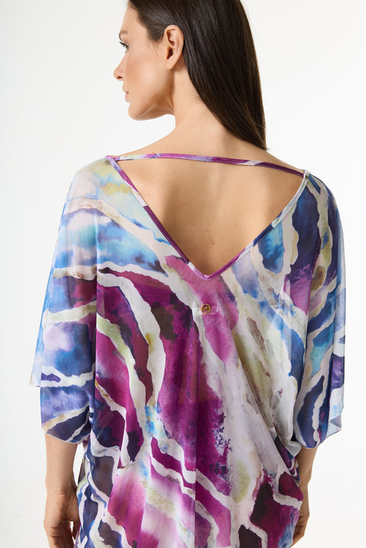 Blusa estampa abstrata colorida em tons de azul, roxo e magenta. A modelo posa de costas, exibindo o decote V profundo, a tira horizontal nas