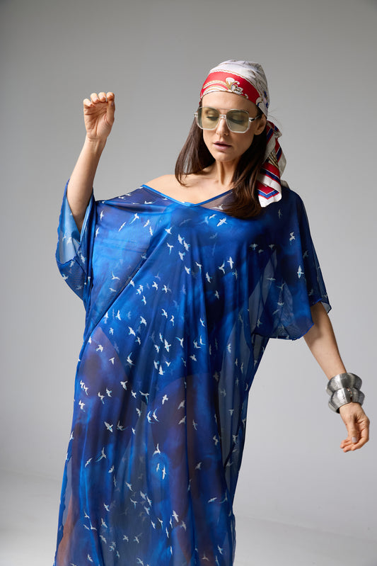 Saída de praia azul com estampa de pássaros brancos. A modelo veste a peça fluida e transparente, com um lenço na cabeça, óculos de sol e