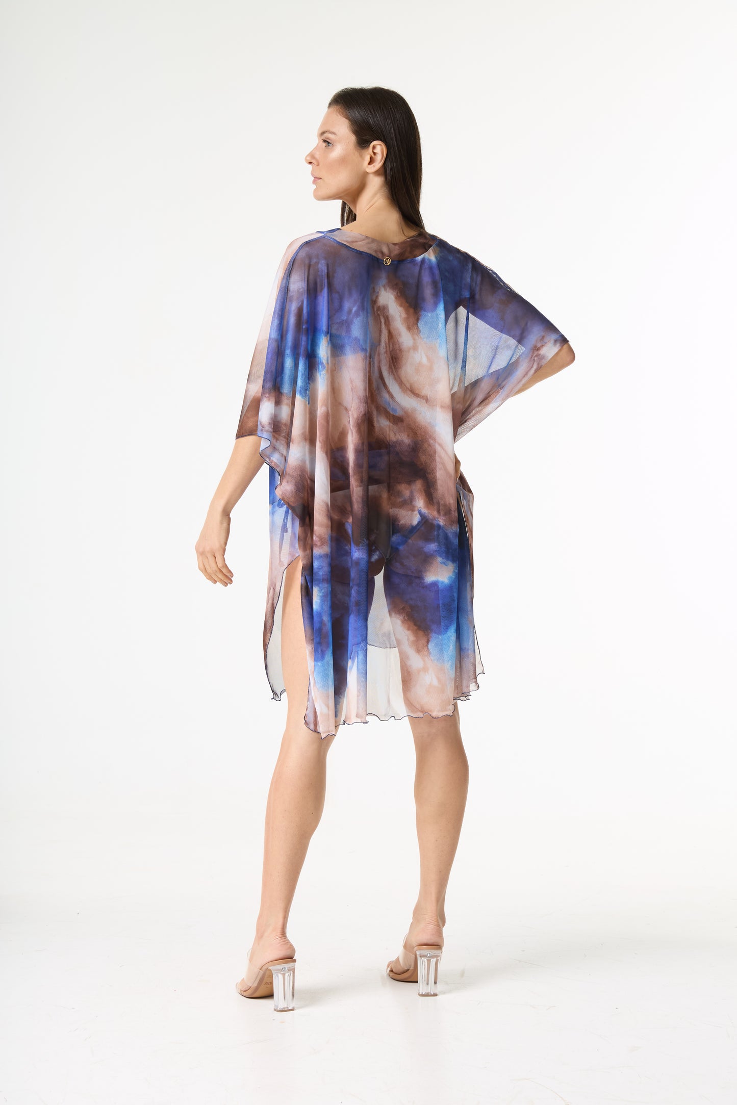 Saída de praia estampa abstrata em tons de azul e marrom. A peça transparente, com caimento fluido, é vista de costas na modelo que vira o rosto para o lado
