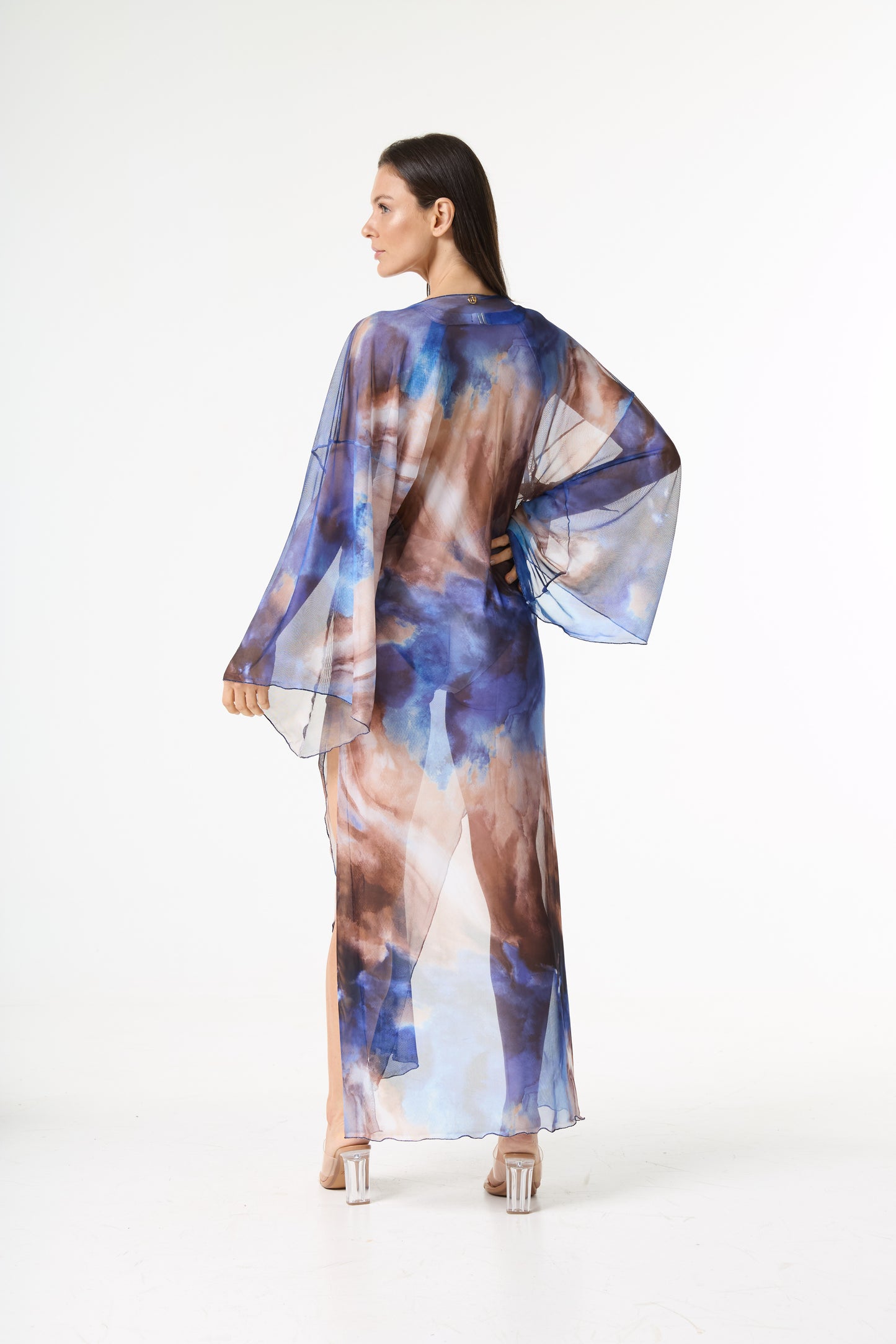 Saída de praia longa transparente estampa aquarela em tons de azul e marrom. A modelo está de costas, olhando por cima do ombro esquerdo, mostrando o caimento fluido da peça em fundo branco.