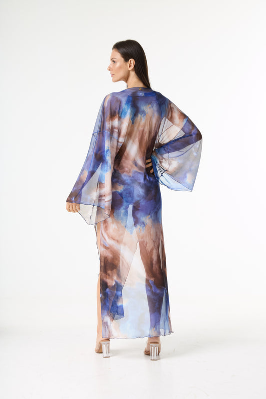 Saída de praia longa transparente estampa aquarela em tons de azul e marrom. A modelo está de costas, olhando por cima do ombro esquerdo, mostrando o caimento fluido da peça em fundo branco.