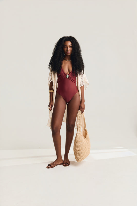 Maiô Siena com decote V e detalhe de pedra amazonita verde, modelo em pose frontal usando saída de praia de crochê off white aberta e bolsa de palha redonda na mão, fundo neutro claro