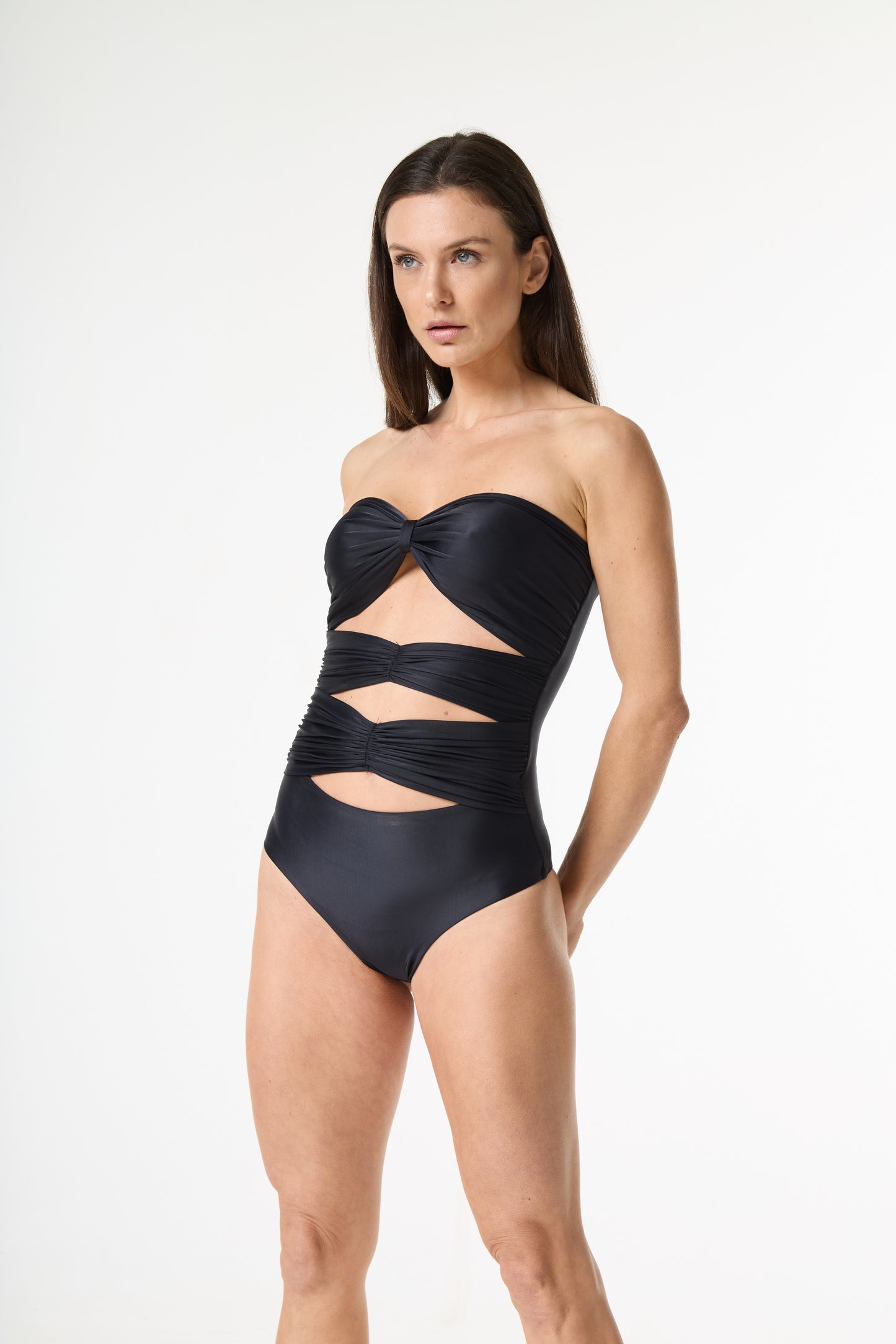 Maiô preto tomara que caia com detalhe de nó no busto e recortes na barriga. A modelo posa de frente, com as mãos atrás das costas, em fundo branco