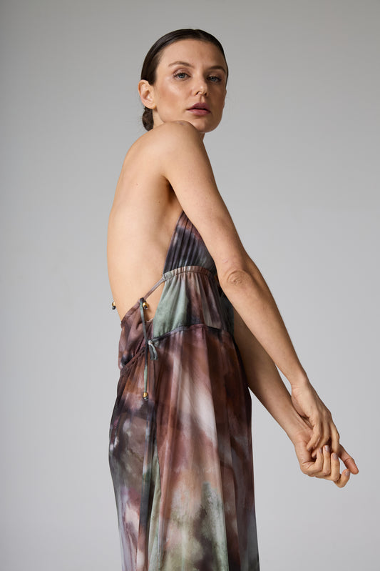 Vestido longo estampa aquarela costas nuas marrom. A modelo está de lado, olhando para a câmera, com o vestido fluido em tons de marrom, verde e cin
