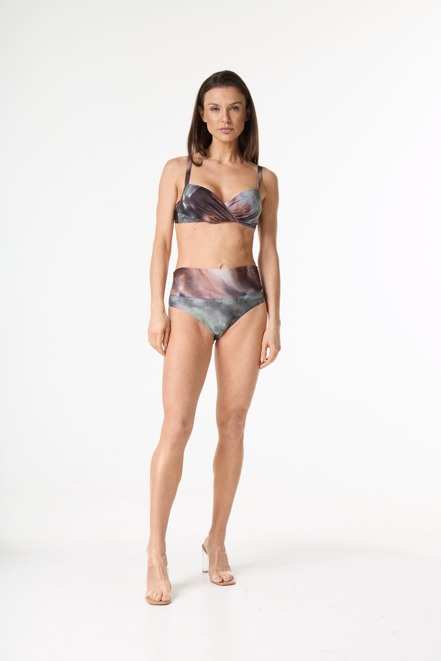 Biquíni estampado de cintura alta com top torcido. A estampa abstrata apresenta tons de marrom, roxo, verde e cinza. Modelo em pé, de frente, em fundo branco, exibindo o caimento da peça e usando sandálias transparentes.