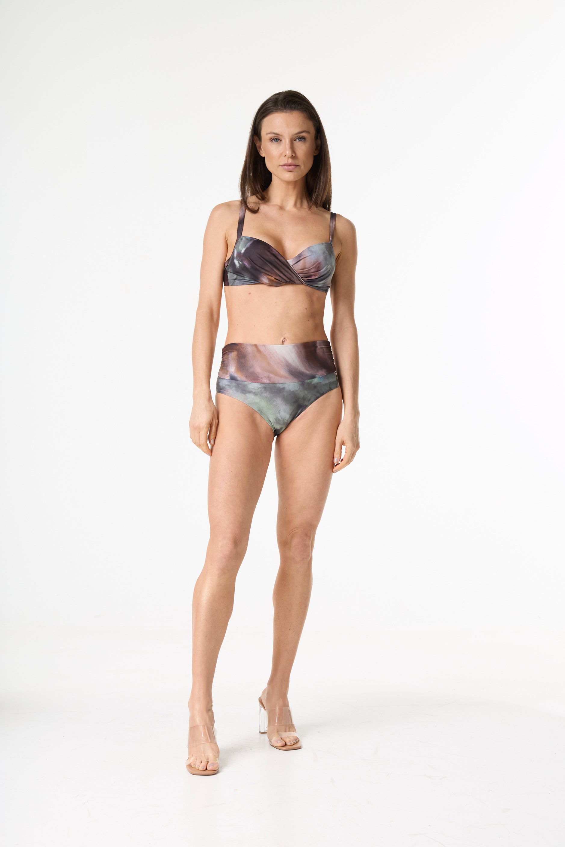Biquíni estampado de cintura alta com top torcido. A estampa abstrata apresenta tons de marrom, roxo, verde e cinza. Modelo em pé, de frente, em fundo branco, exibindo o caimento da peça e usando sandálias transparentes.