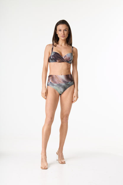 Biquíni estampado de cintura alta com top torcido. A estampa abstrata apresenta tons de marrom, roxo, verde e cinza. Modelo em pé, de frente, em fundo branco, exibindo o caimento da peça e usando sandálias transparentes.