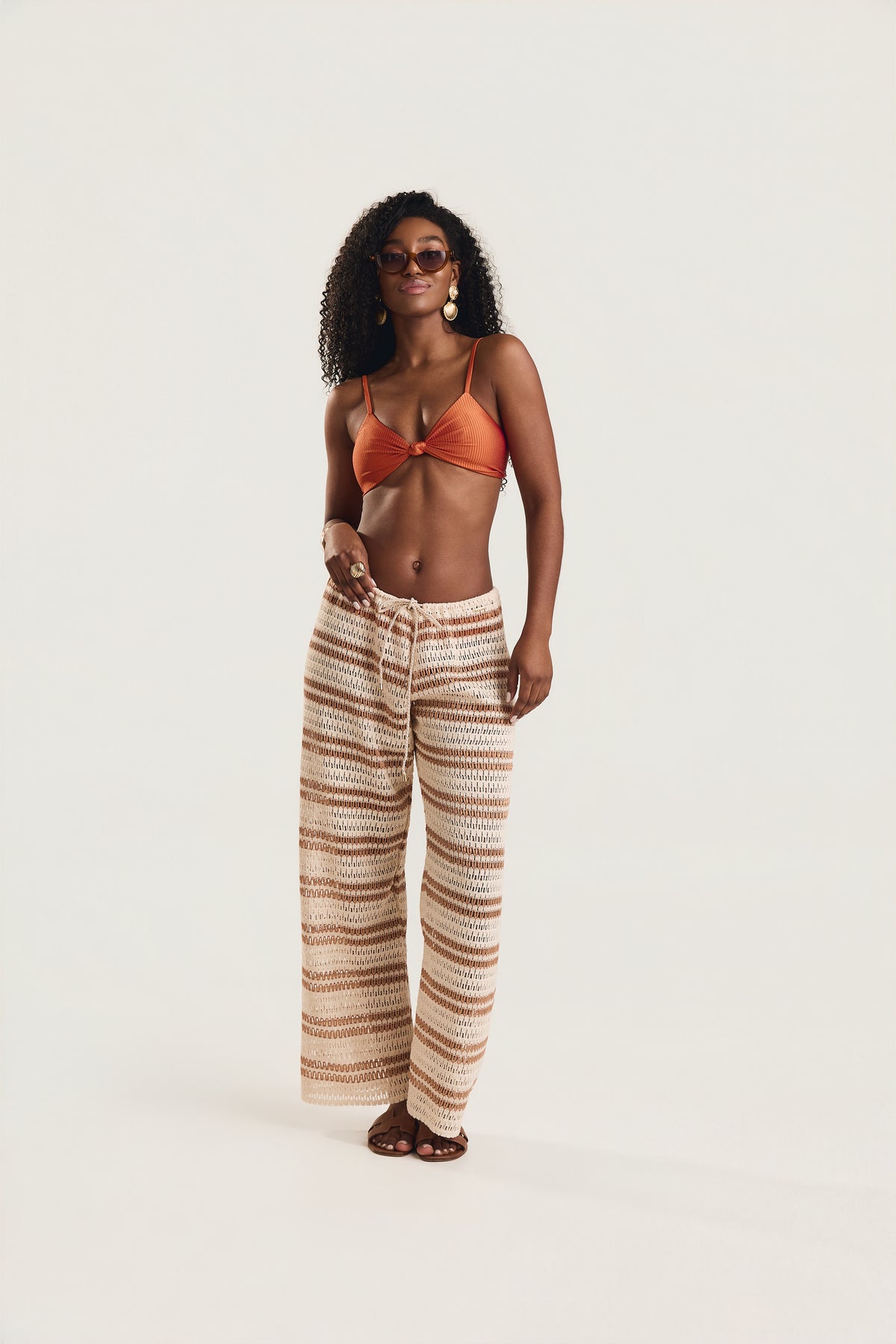 Calça pantalona de crochê listrada em off white e caramelo, modelo em postura frontal com uma mão na cintura e óculos de sol