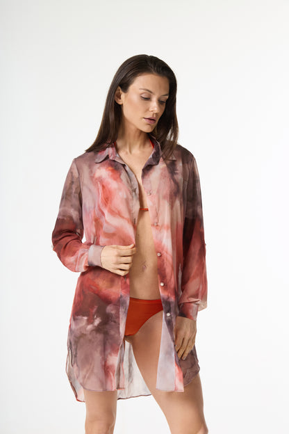 Saída de praia estampa aquarela. A modelo usa a camisa longa e transparente com padrões em tons de vermelho, rosa e marrom, aberta sobre um biquíni laranja, em fundo branco. Ela segura a barra da camisa e olha para baixo.