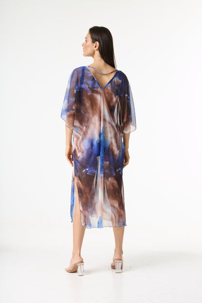 Saída de praia kaftan estampa tie dye. A modelo está de costas, olhando para o lado, exibindo o caimento fluido da peça transparente em tons de azul e marrom