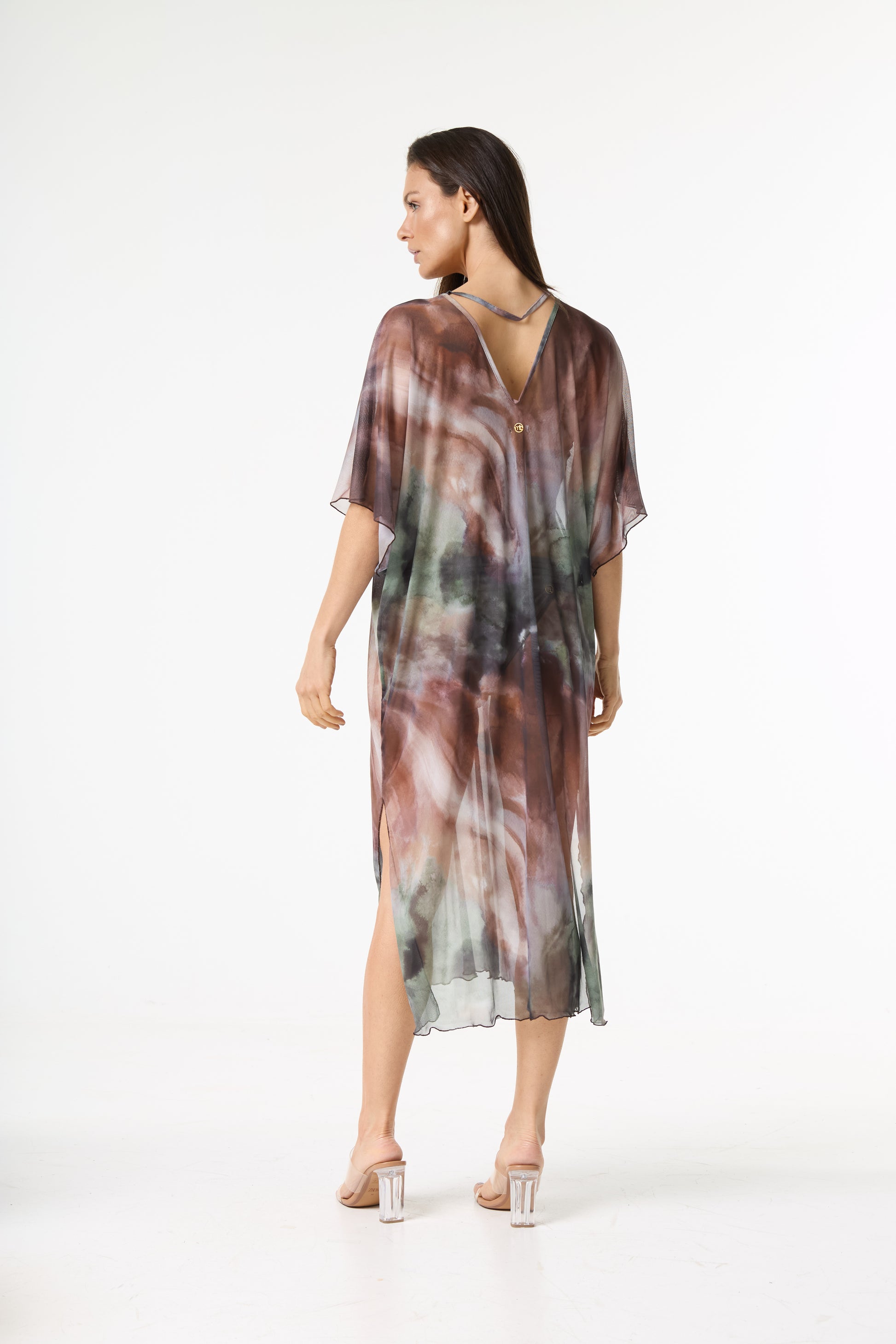 Vestido kaftan longo estampado. Modelo de costas, olhando por cima do ombro esquerdo, veste a peça transparente com estampa abstrata em tons de marrom, verde e toques de cinza