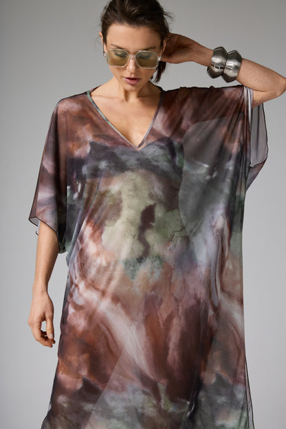 Túnica transparente com estampa aquarelada em tons terrosos de marrom, verde e cinza. A modelo posa de frente, com óculos de sol e tocando a cabeça, exibindo o caimento fluido e o decote V da peça.