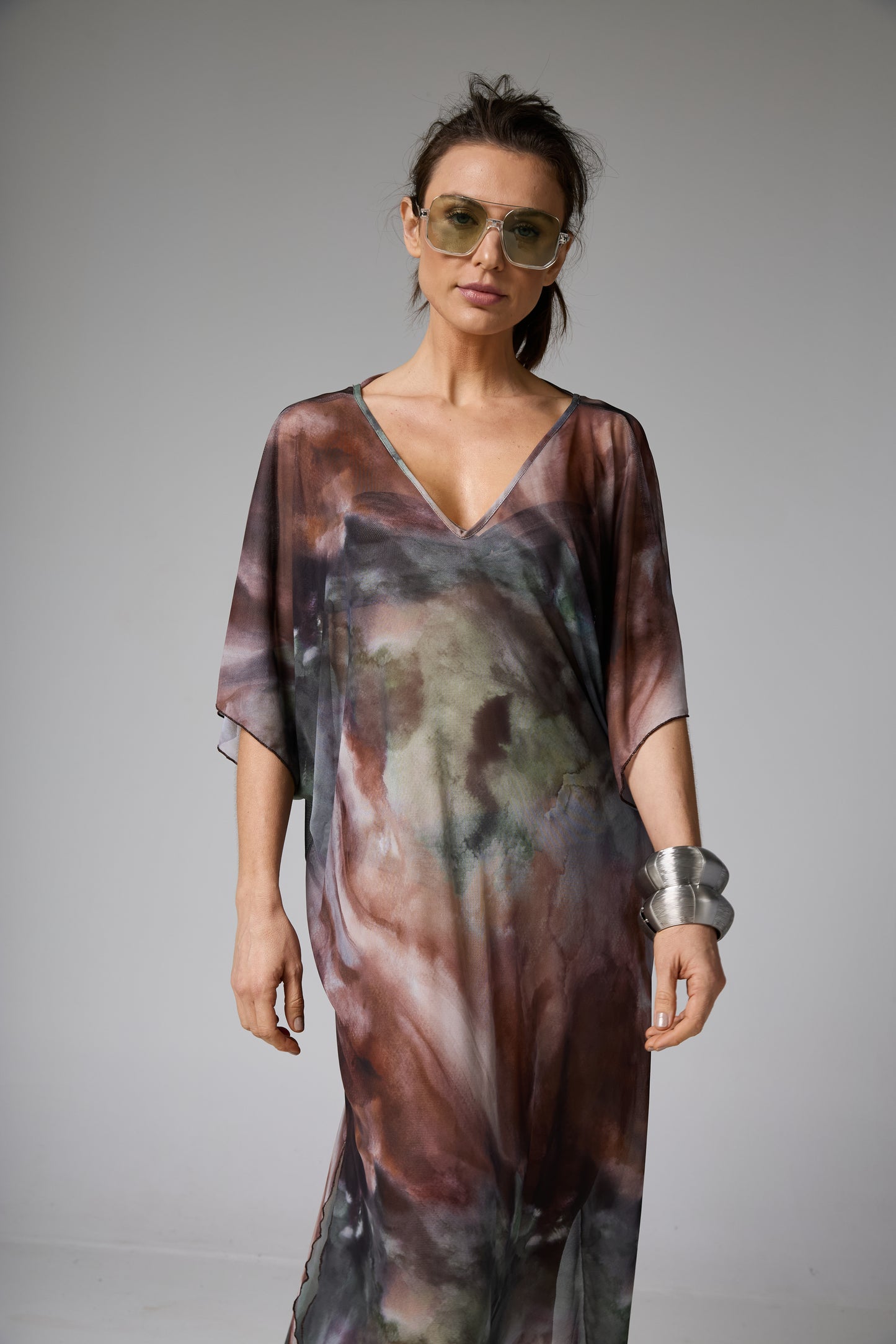 Caftan longo de tecido fluido com decote V e estampa abstrata em tons de marrom, verde e cinza. A modelo usa óculos de sol e pulseiras volumosas