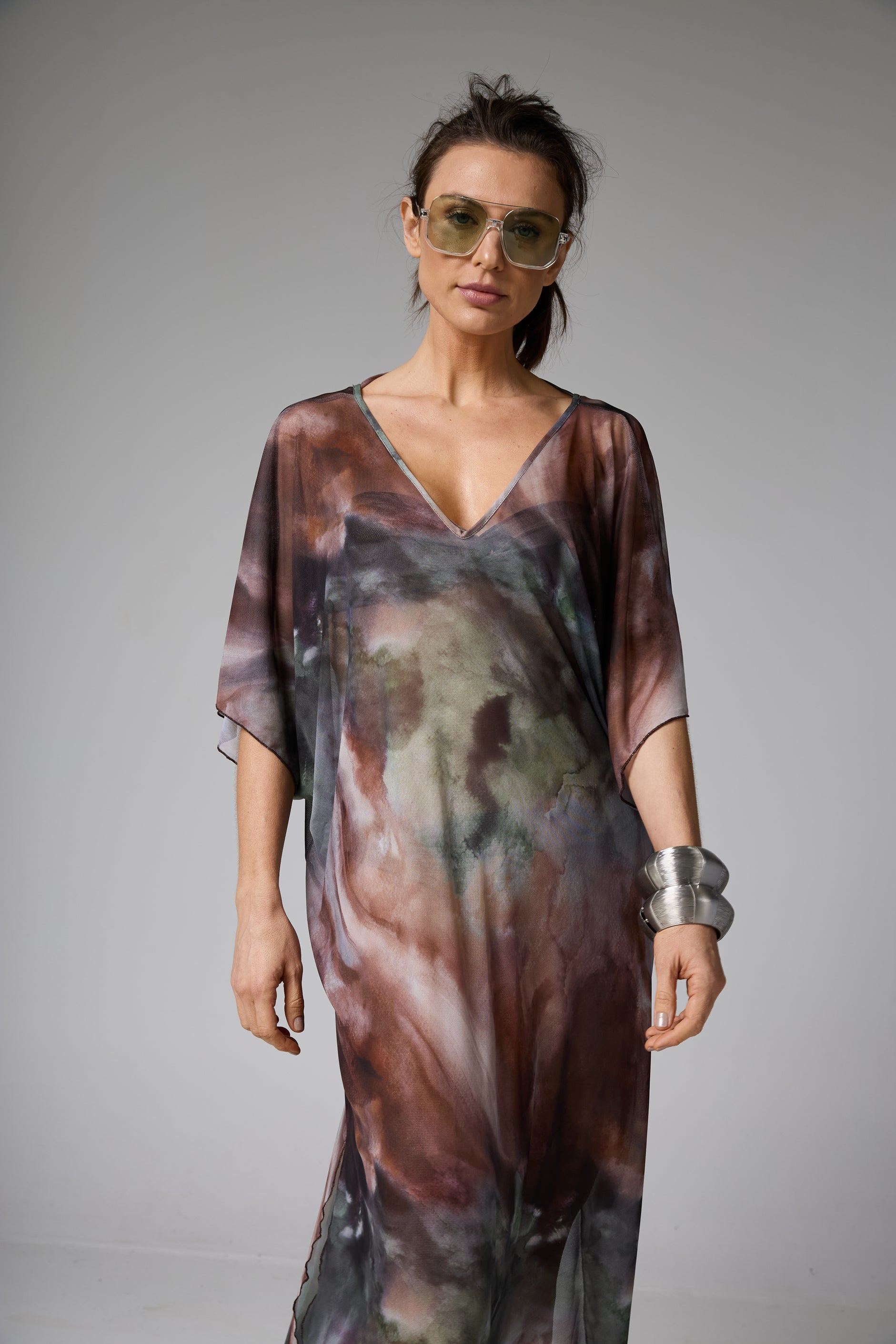Caftan longo de tecido fluido com decote V e estampa abstrata em tons de marrom, verde e cinza. A modelo usa óculos de sol e pulseiras volumosas