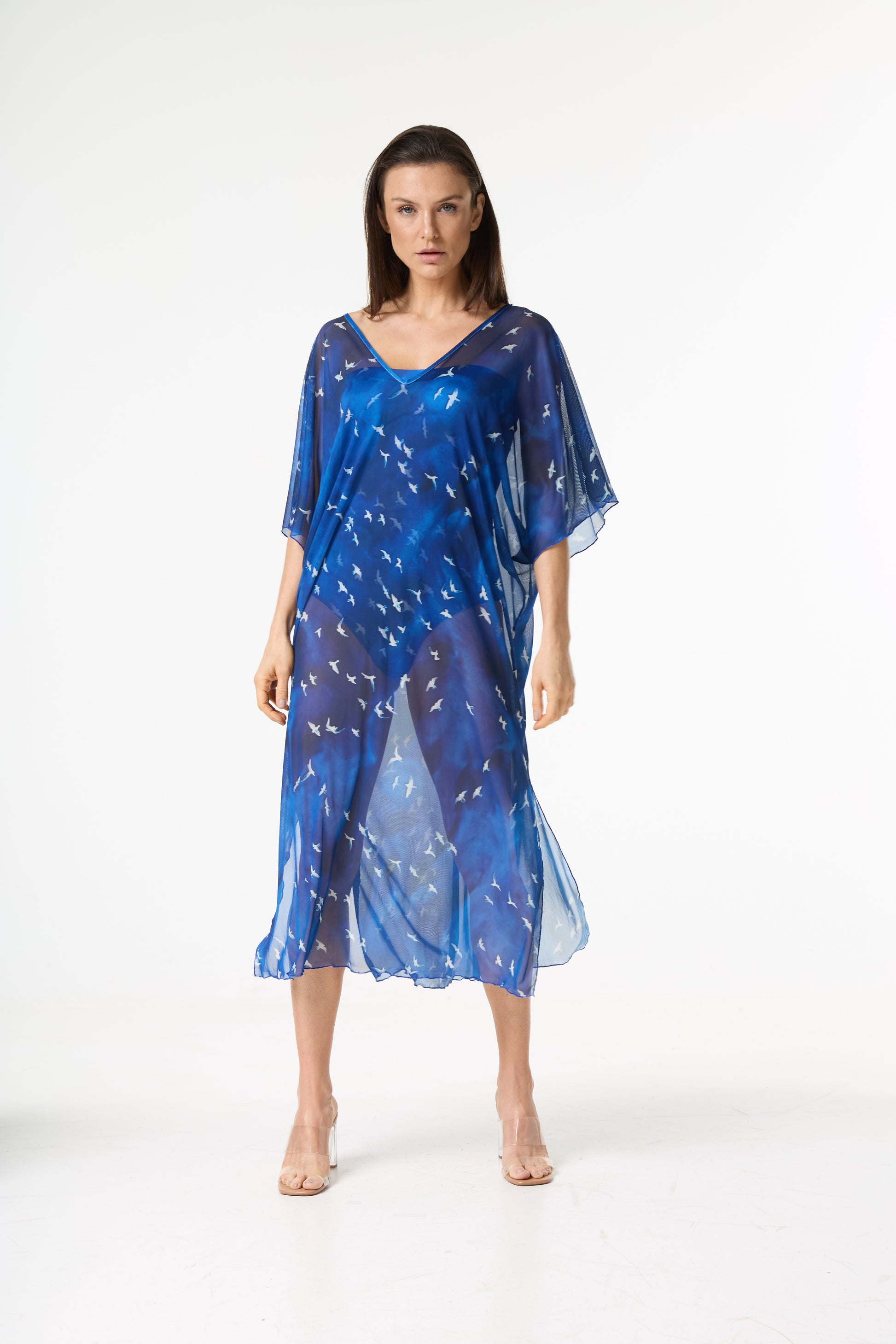 Saída de praia kaftan azul com estampa de pássaros brancos. A modelo está de pé, de frente para a câmera, em fundo branco, realçando a transparência e o caimento da peça sobre o biquíni azul.
