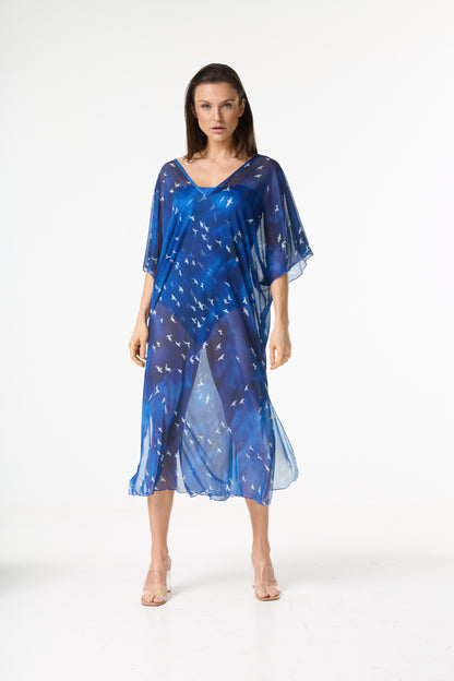 Saída de praia kaftan azul com estampa de pássaros brancos. A modelo está de pé, de frente para a câmera, em fundo branco, realçando a transparência e o caimento da peça sobre o biquíni azul.