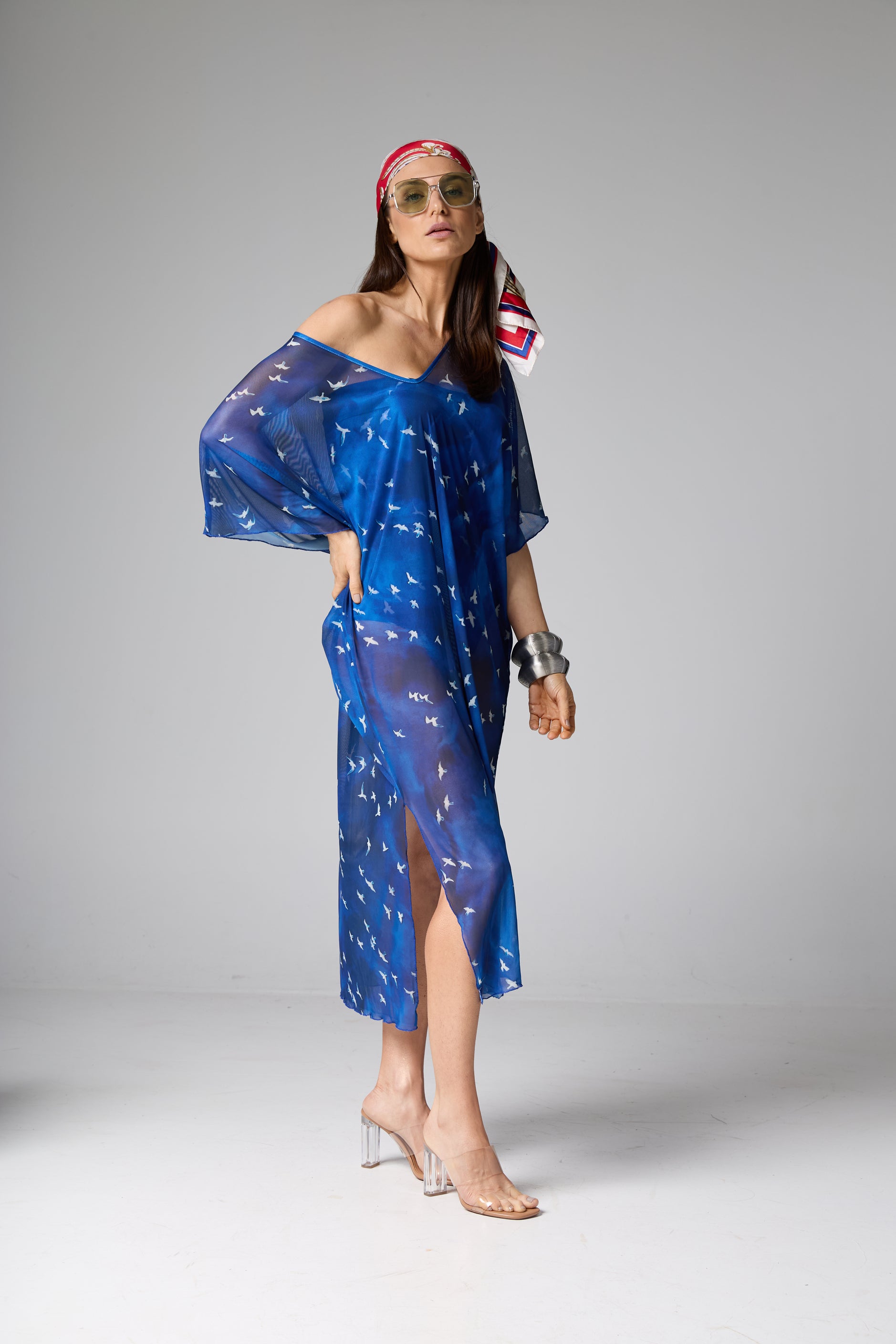 Vestido kaftan azul midi transparente com estampa de pássaros brancos voando. A modelo está de pé com a mão na cintura, usando óculos de sol, lenço na cabeça e pulseira grande.
