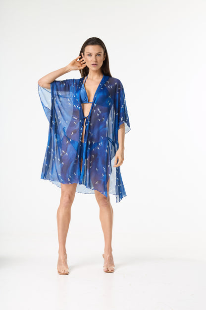 Saída de praia kimono azul com estampa de pássaros brancos. A modelo posa de frente em fundo branco, com a mão no cabelo, mostrando o caimento leve da peça sobre