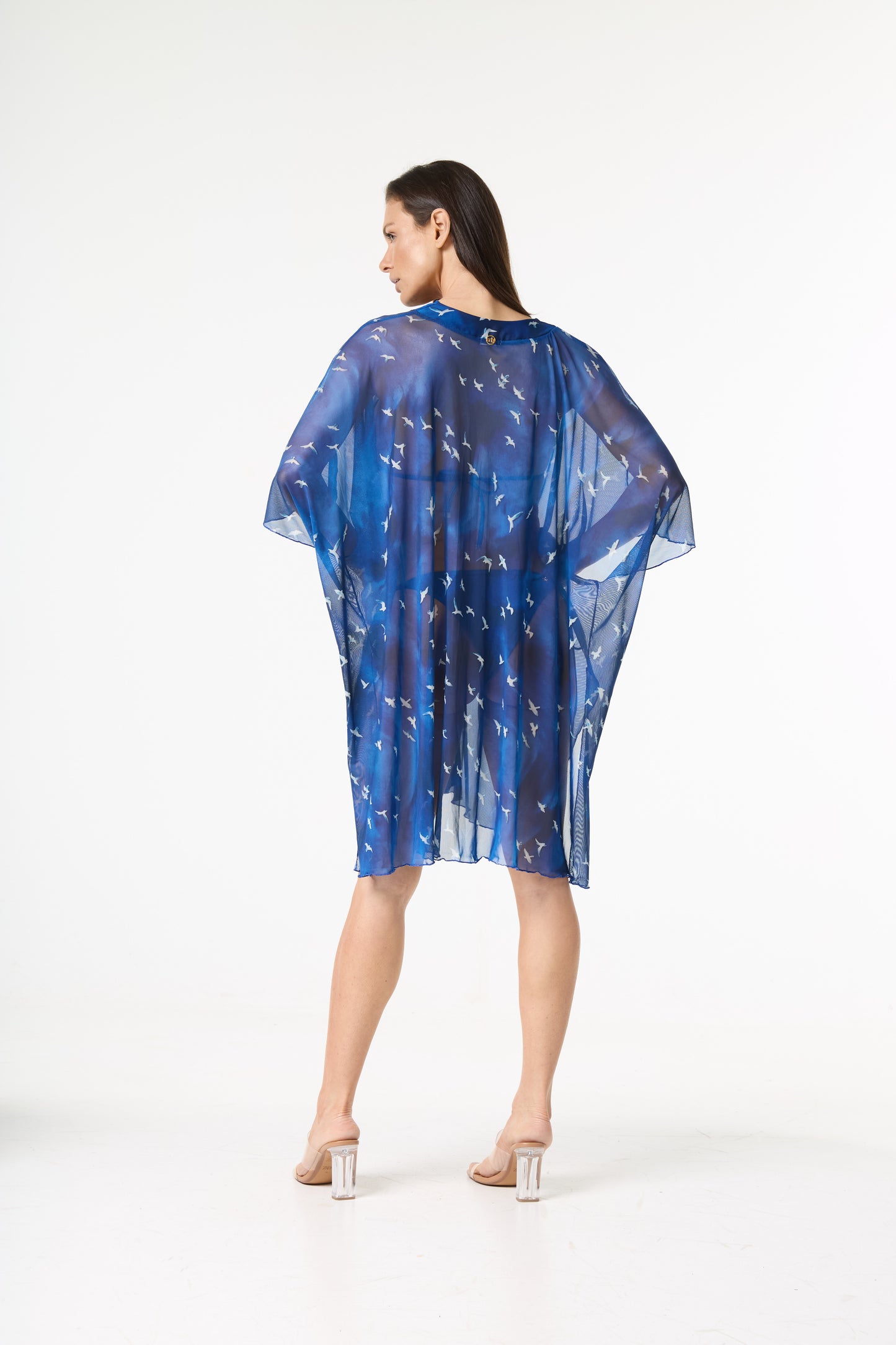 Saída de praia kaftan azul com estampa de pássaros. A modelo está de costas para a câmera, com o rosto virado para o lado esquerdo e as mãos na cintura,