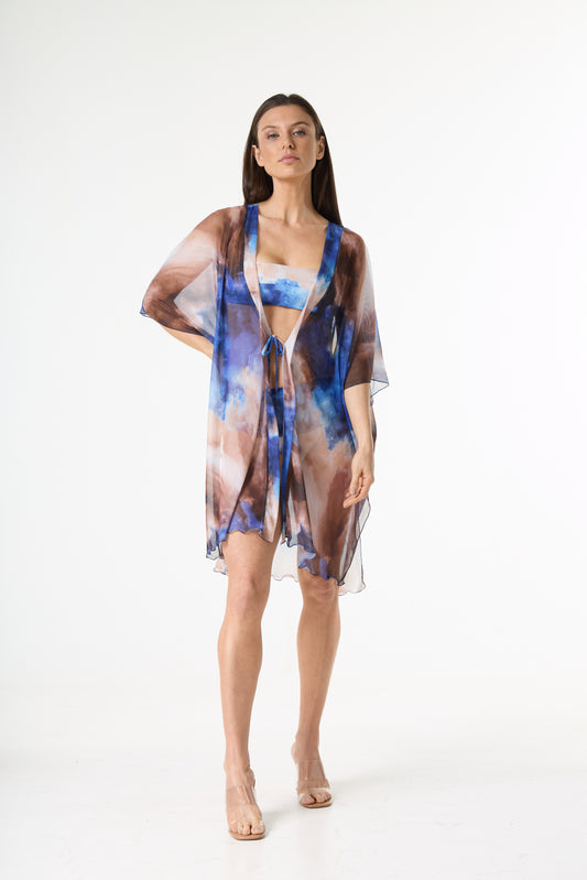 Saída de praia quimono com estampa abstrata em tons de azul e marrom. A modelo está de frente, de corpo inteiro, em estúdio branco, exibindo a peça que inclui um quimono transparente de mangas largas e um biquíni bandeau com a mesma estampa.