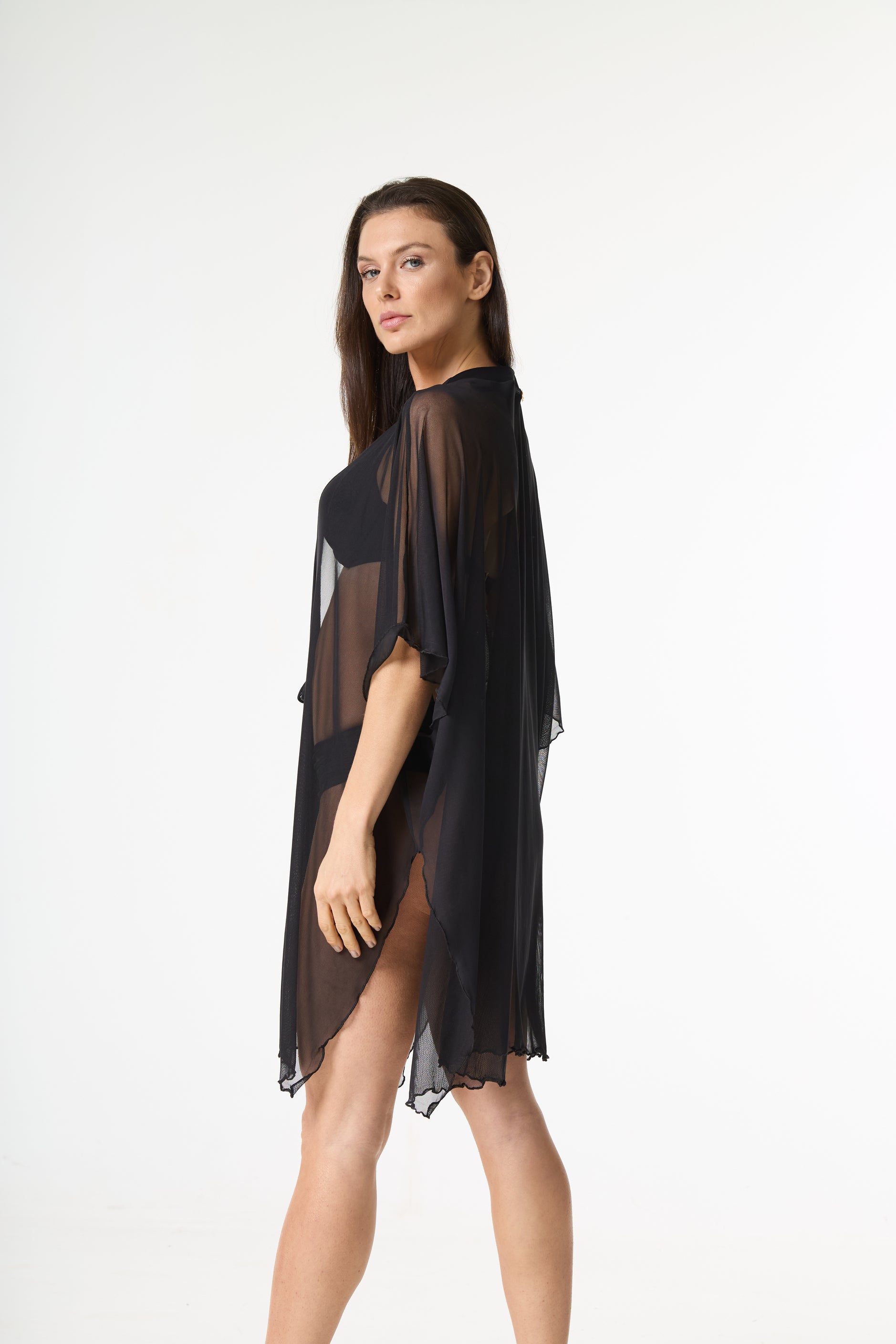 Saída de praia kaftan preta transparente. A modelo usa a peça leve e fluida sobre um biquíni preto, posando de lado e olhando para a câmera em um fundo