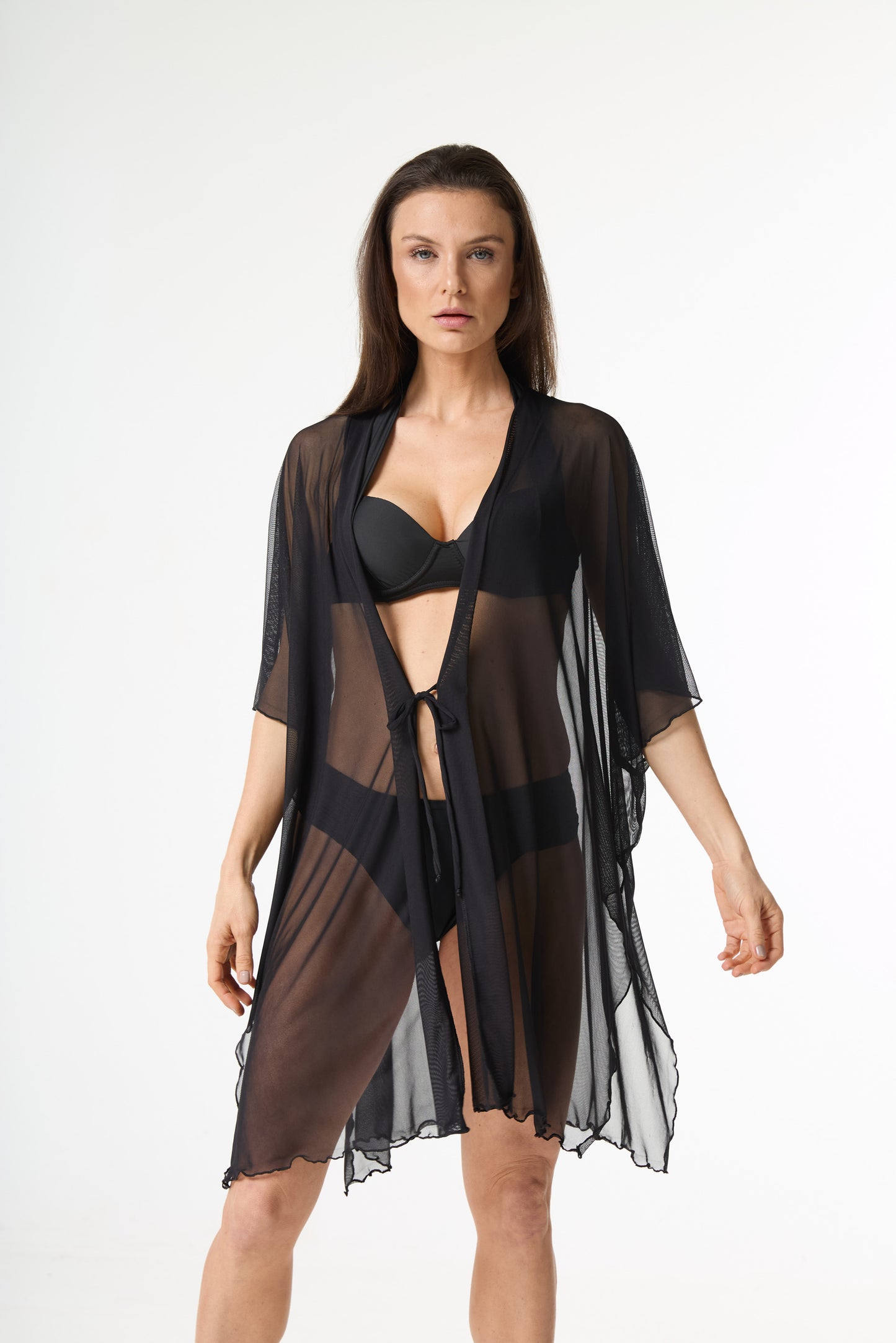 Saída de praia tule preta. Modelo veste a peça preta translúcida estilo quimono com amarra na cintura sobre biquíni preto, posando de frente em fundo branco