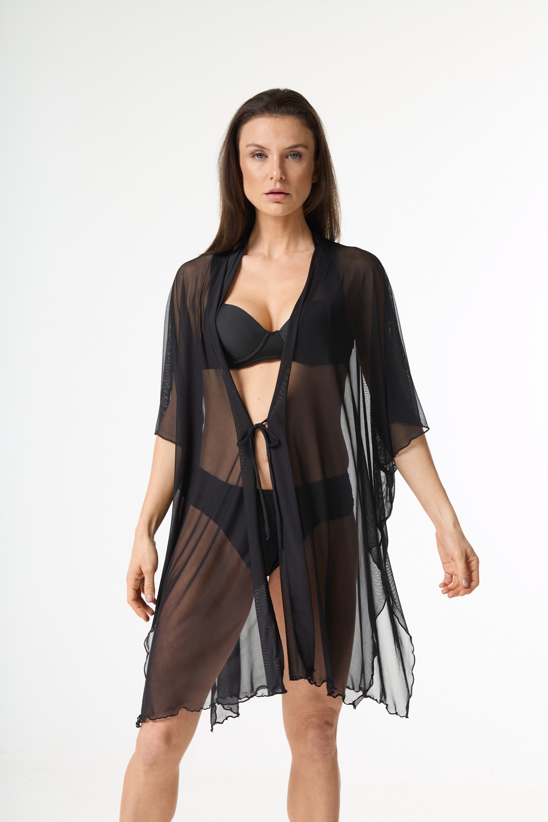 Saída de praia tule preta. Modelo veste a peça preta translúcida estilo quimono com amarra na cintura sobre biquíni preto, posando de frente em fundo branco