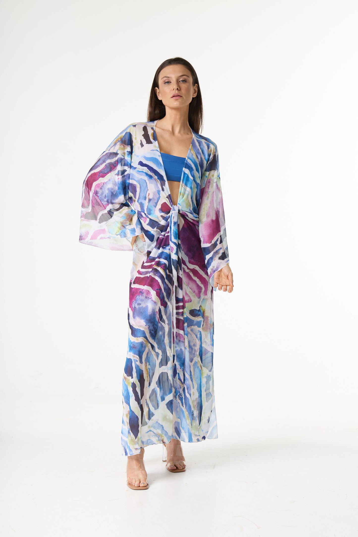 Saída de praia longa com estampa abstrata em tons de azul, roxo e rosa, com amarração frontal. A modelo posa de frente em estúdio branco, destacando o caimento leve da peça.