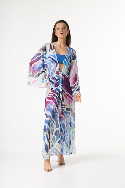 Saída de praia longa com estampa abstrata em tons de azul, roxo e rosa, com amarração frontal. A modelo posa de frente em estúdio branco, destacando o caimento leve da peça.