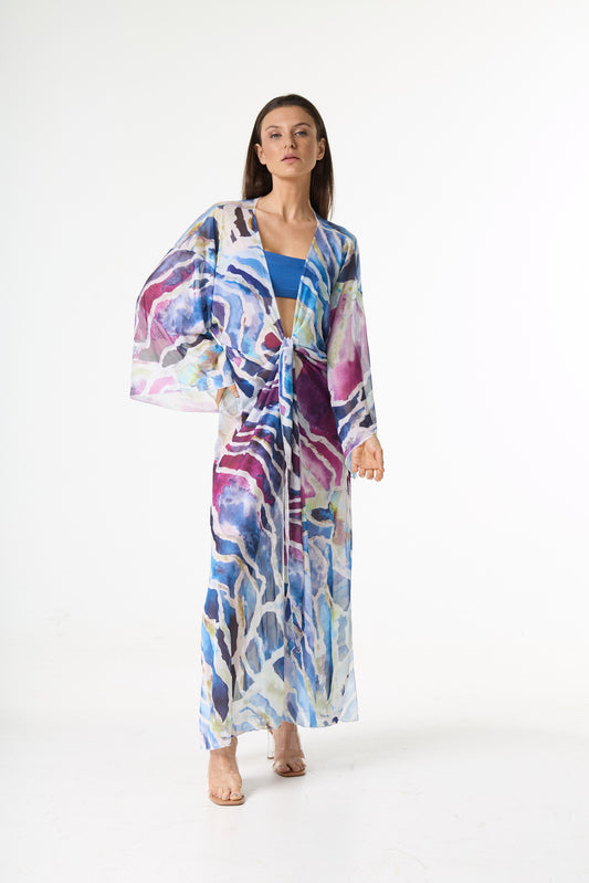 Saída de praia longa com estampa abstrata em tons de azul, roxo e rosa, com amarração frontal. A modelo posa de frente em estúdio branco, destacando o caimento leve da peça.