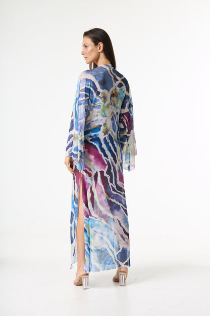 Saída de praia longa com estampa abstrata em tons de azul, roxo e verde. A modelo está de costas para a câmera, com o rosto virado para o lado es