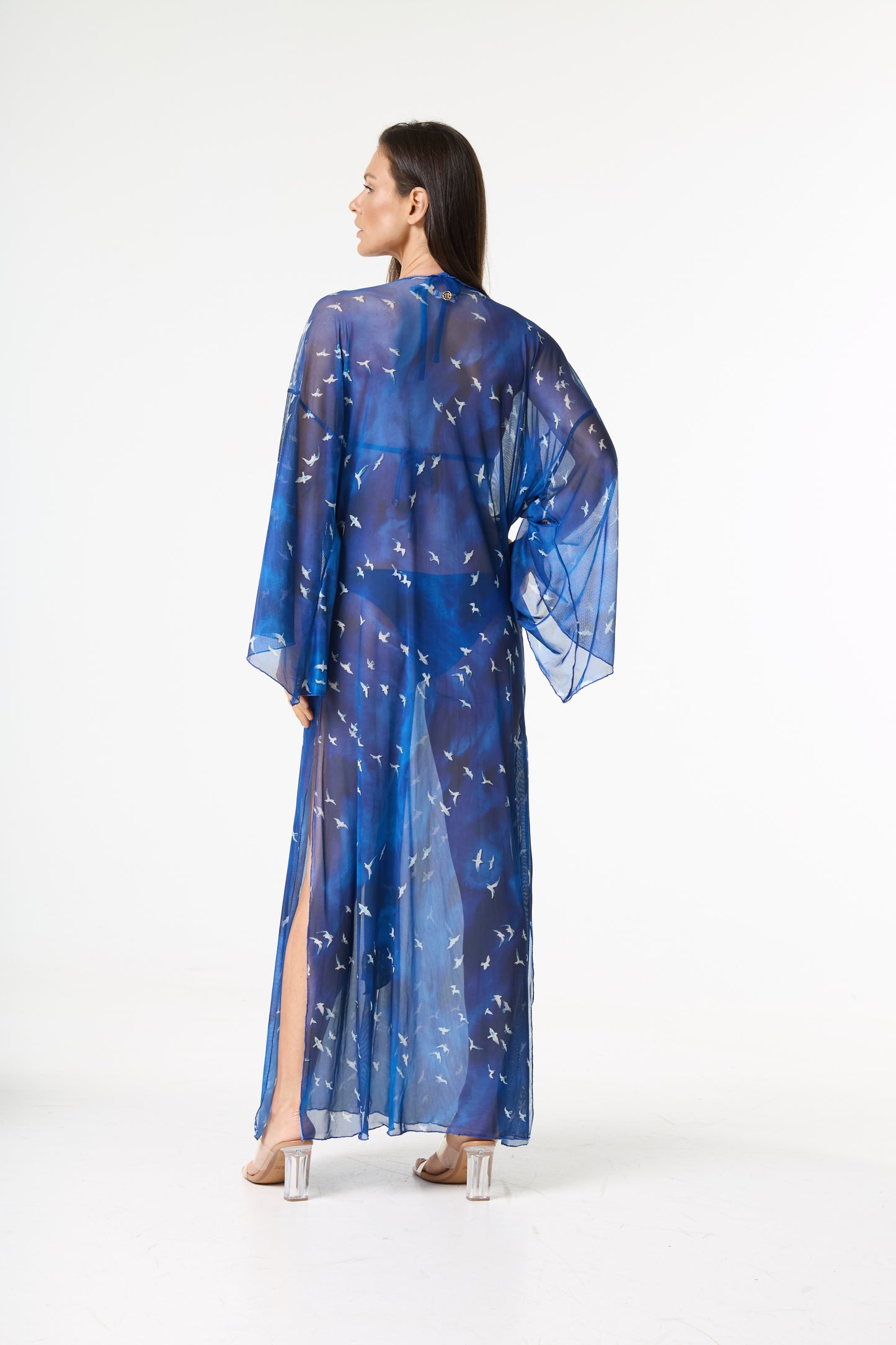 Saída de praia longa azul transparente com estampa de pássaros brancos. A modelo, de costas e virada de lado, exibe o caimento fluido e a fenda da peça sobre um maiô escuro, usando sandálias de salto transparente.