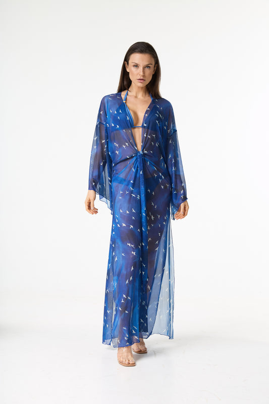 Saída de praia longa azul estampa pássaros. Modelo de frente em fundo branco, com a peça de tecido translúcido amarrada na cintura, exibindo o decote V e mangas amplas.