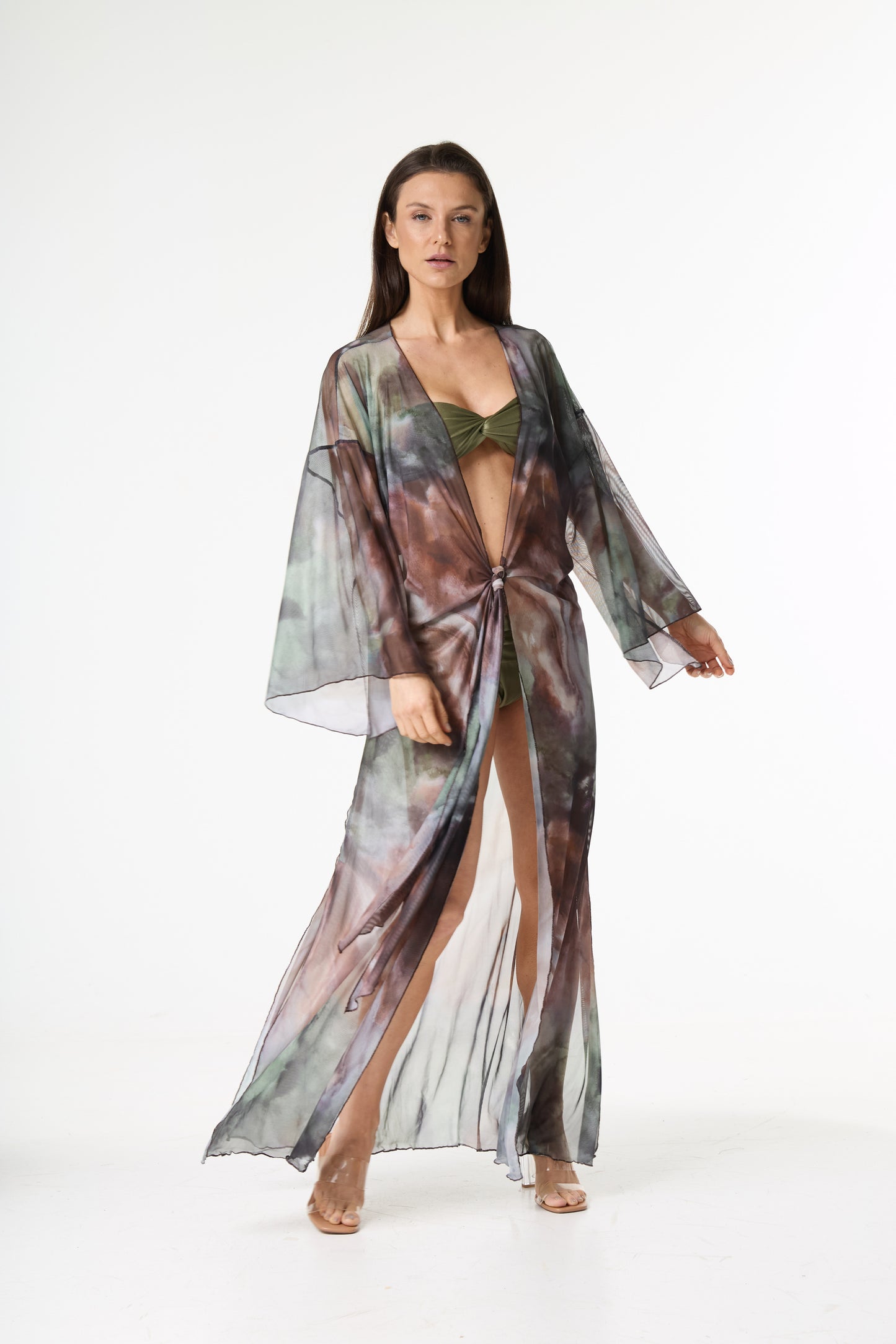Saída de praia longa com estampa abstrata em tons de marrom, verde e cinza. A modelo está de frente, exibindo o caimento da peça transparente, amarrada na cintura, com fenda e mangas largas, sobre um biquíni verde em fundo branco.