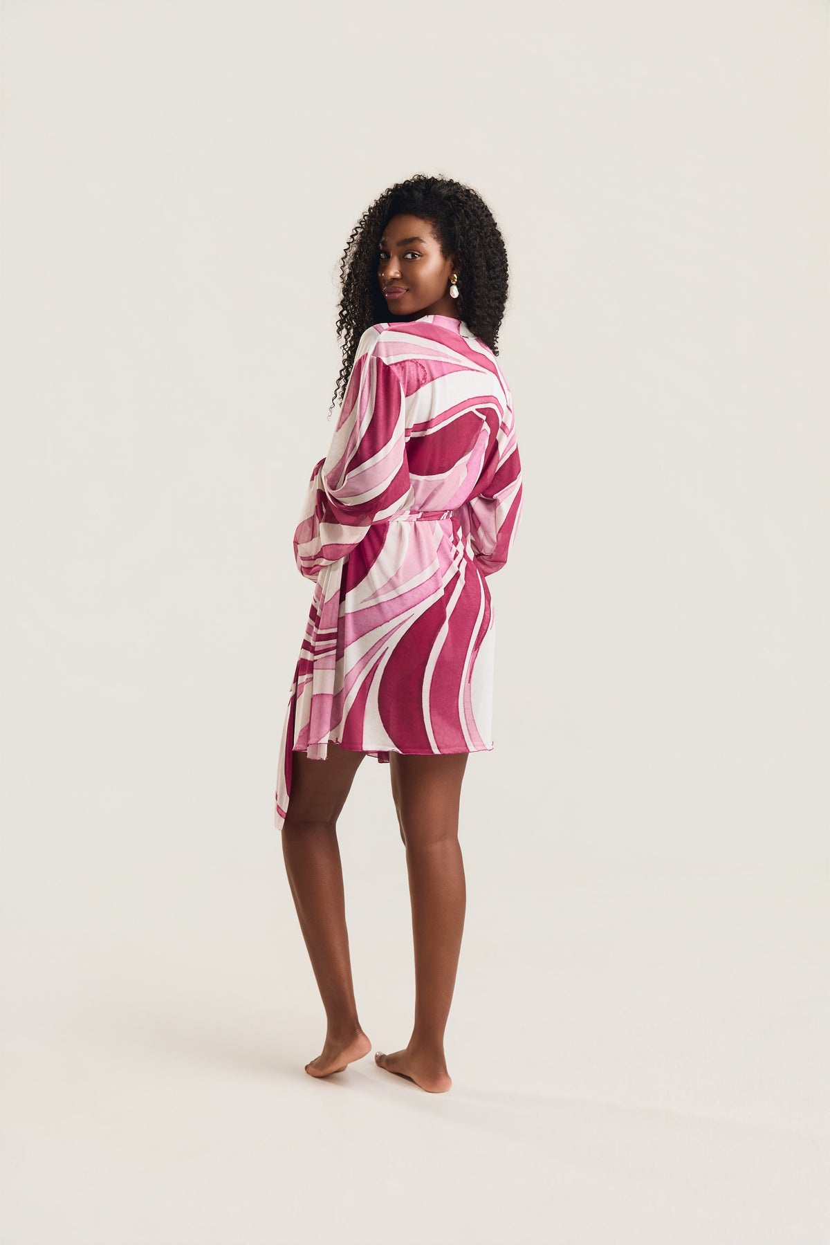 Kimono de manga longa com estampa geo rosa visto de costas, modelo em posição de três quartos olhando por cima do ombro, descalça
