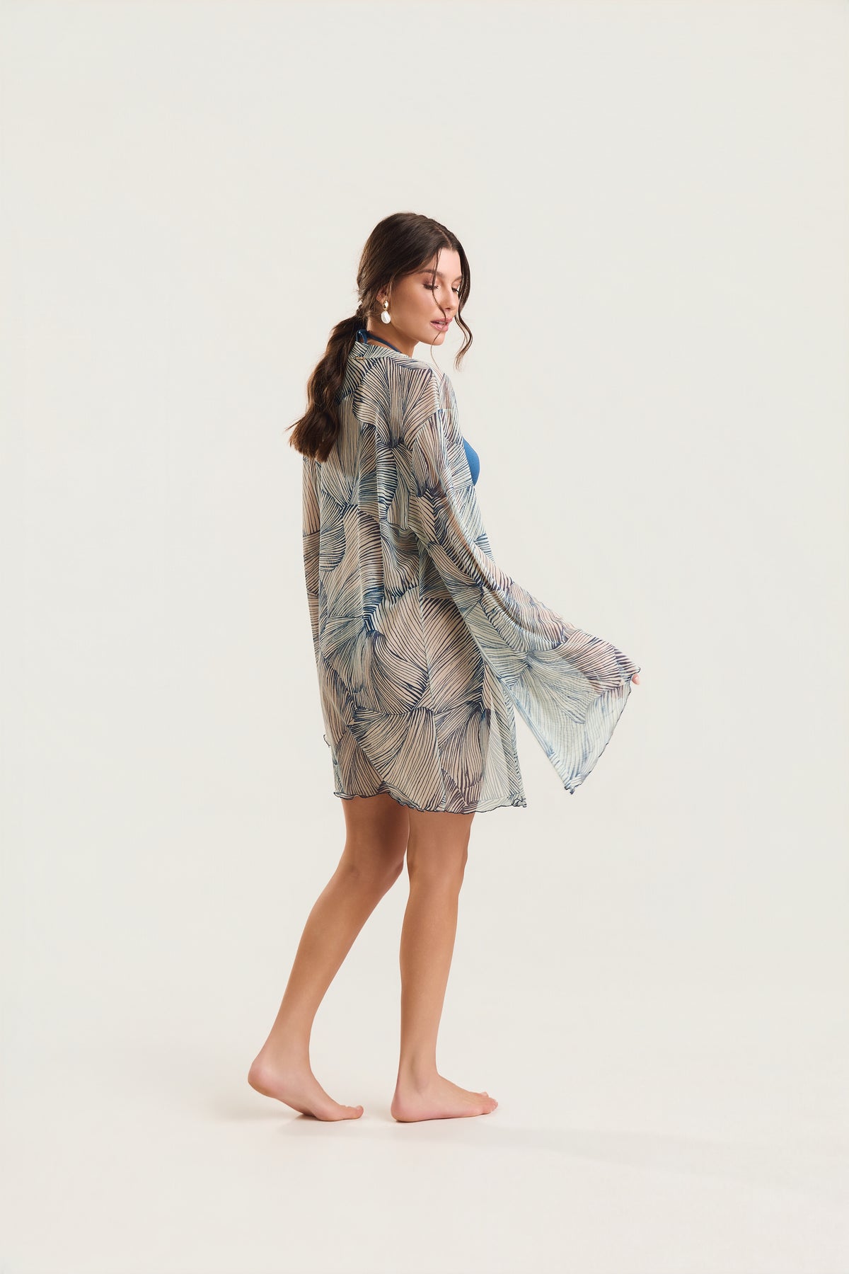 Kimono de manga longa com estampa de conchas azuis visto de costas, modelo em posição de perfil caminhando, com o tecido fluido ao vento
