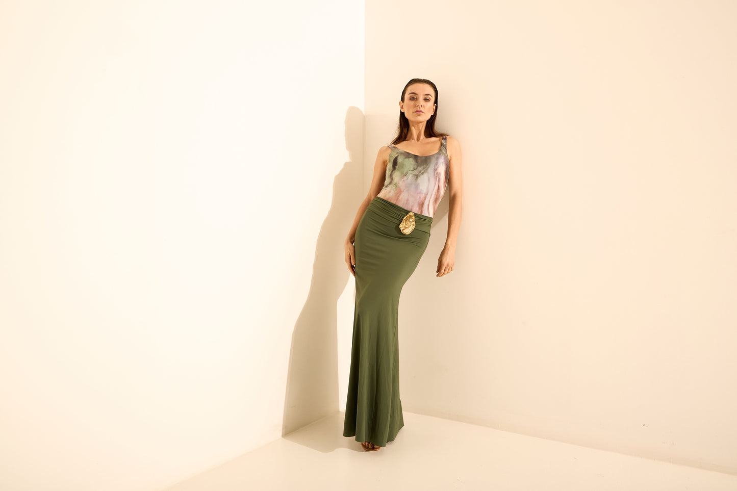 Saia longa verde oliva com cinto dourado e regata estampada. A modelo está de frente, encostada na parede, exibindo o caimento justo das peças.