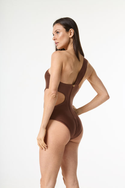 Body maiô um ombro marrom com recortes laterais e emblema dourado. A modelo posa de costas, olhando por cima do ombro, realçando o ajuste da peça e os detalhes do design.