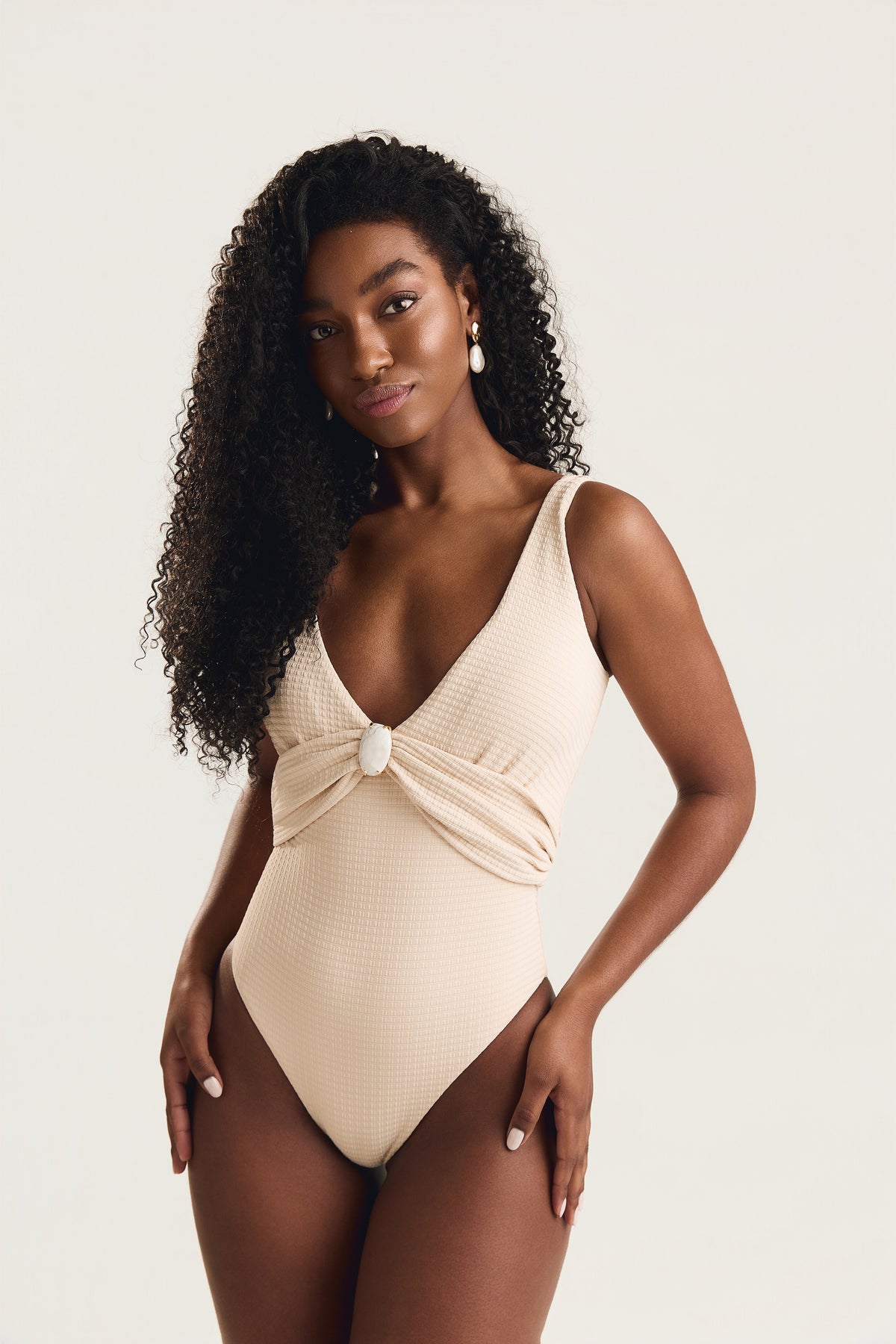 Maiô off white textura com decote V drapeado e pedra howlita branca no centro, modelo em pose frontal com mãos no quadril e cabelos cacheados soltos, corpo inteiro, fundo claro