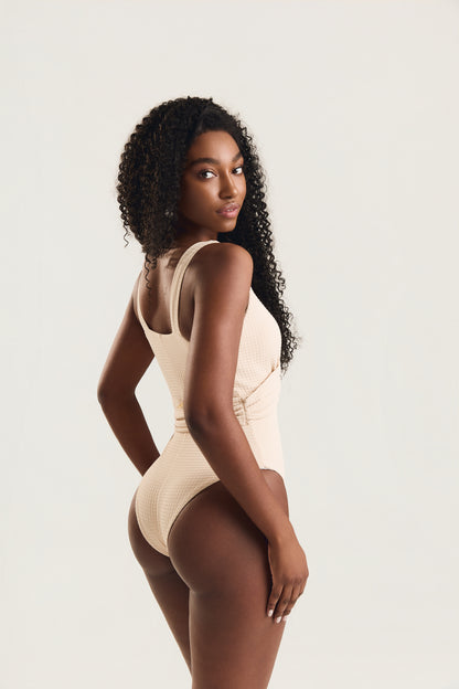 Maiô off white textura com alças largas e corte nas costas, modelo em pose de costas com leve giro do rosto para o lado, mão no quadril evidenciando o caimento e o recorte traseiro, fundo claro