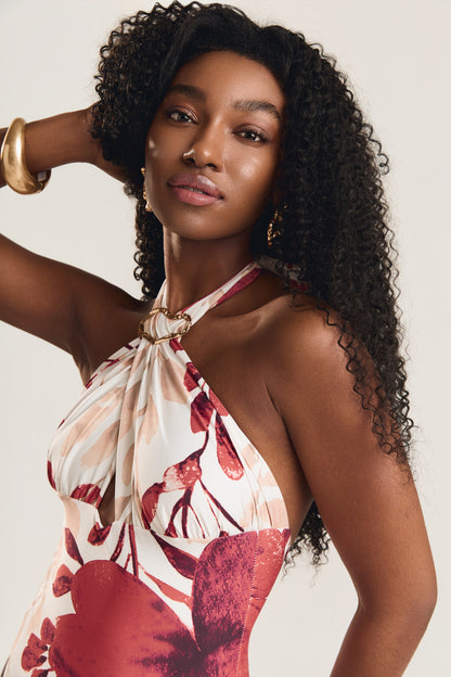 Close no busto da modelo com braço levantado atrás da cabeça e olhar lateral, destacando o decote halter cruzado com argola dourada do maiô com estampa floral vermelho