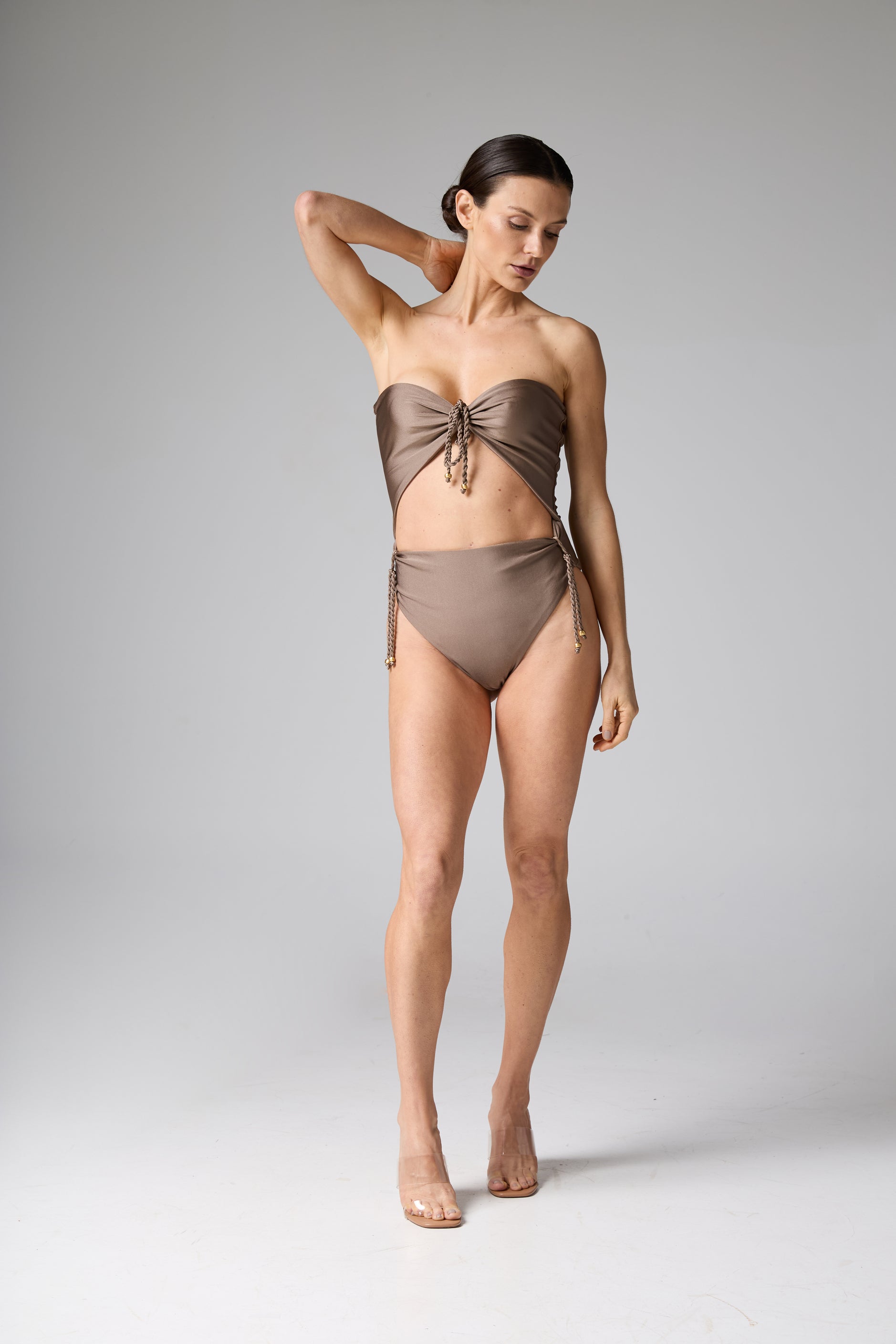 Maiô monokini tomara que caia recortes trançados bronze. A modelo posa de pé, com uma mão na nuca, exibindo a modelagem com