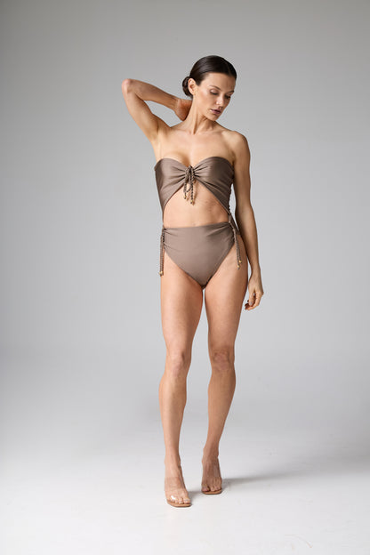 Maiô monokini tomara que caia recortes trançados bronze. A modelo posa de pé, com uma mão na nuca, exibindo a modelagem com