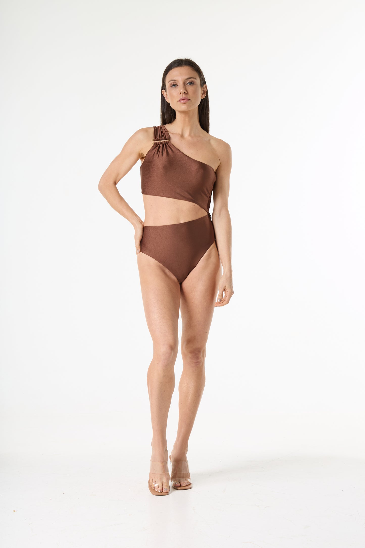 Maiô bronze de um ombro só com recorte na cintura e detalhe de argola na alça. Modelo posa de frente, com a mão na cintura, em fundo branco.