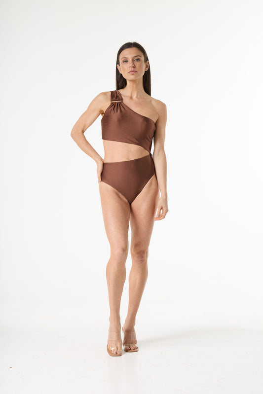 Maiô bronze de um ombro só com recorte na cintura e detalhe de argola na alça. Modelo posa de frente, com a mão na cintura, em fundo branco.