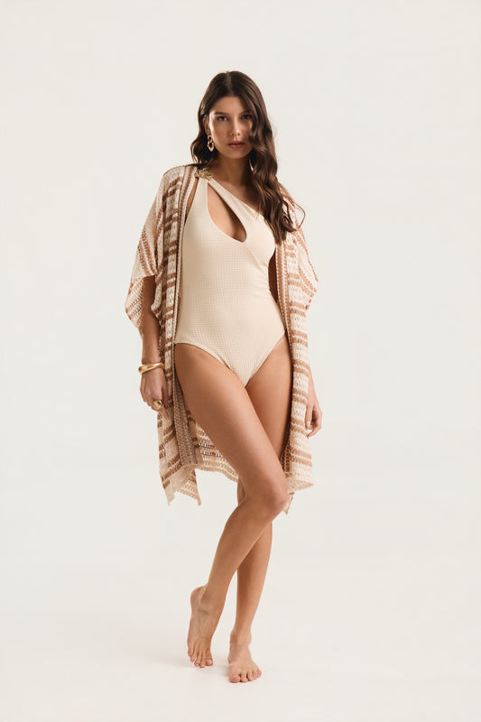 Maiô Um Ombro Metal Amassado off white textura, modelo em pose frontal com perna cruzada à frente, usando saída de praia kimono estampada em tons terrosos, fundo neutro claro.

