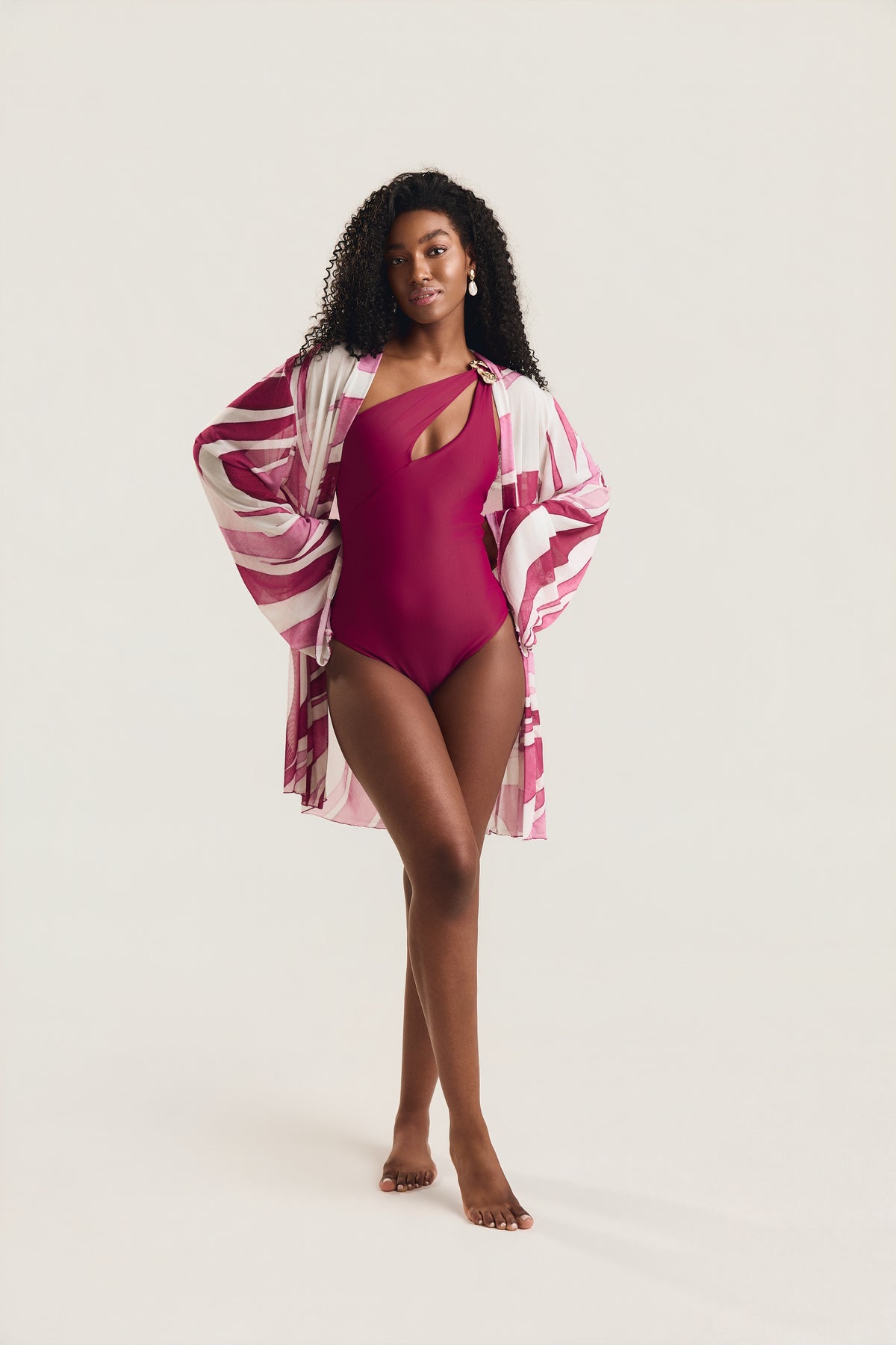 Maiô Um Ombro Metal Amassado carmim, modelo em pose frontal com pernas cruzadas usando saída de praia kimono estampada em geo rosa e branco, braço estendido para o lado, fundo neutro claro.

