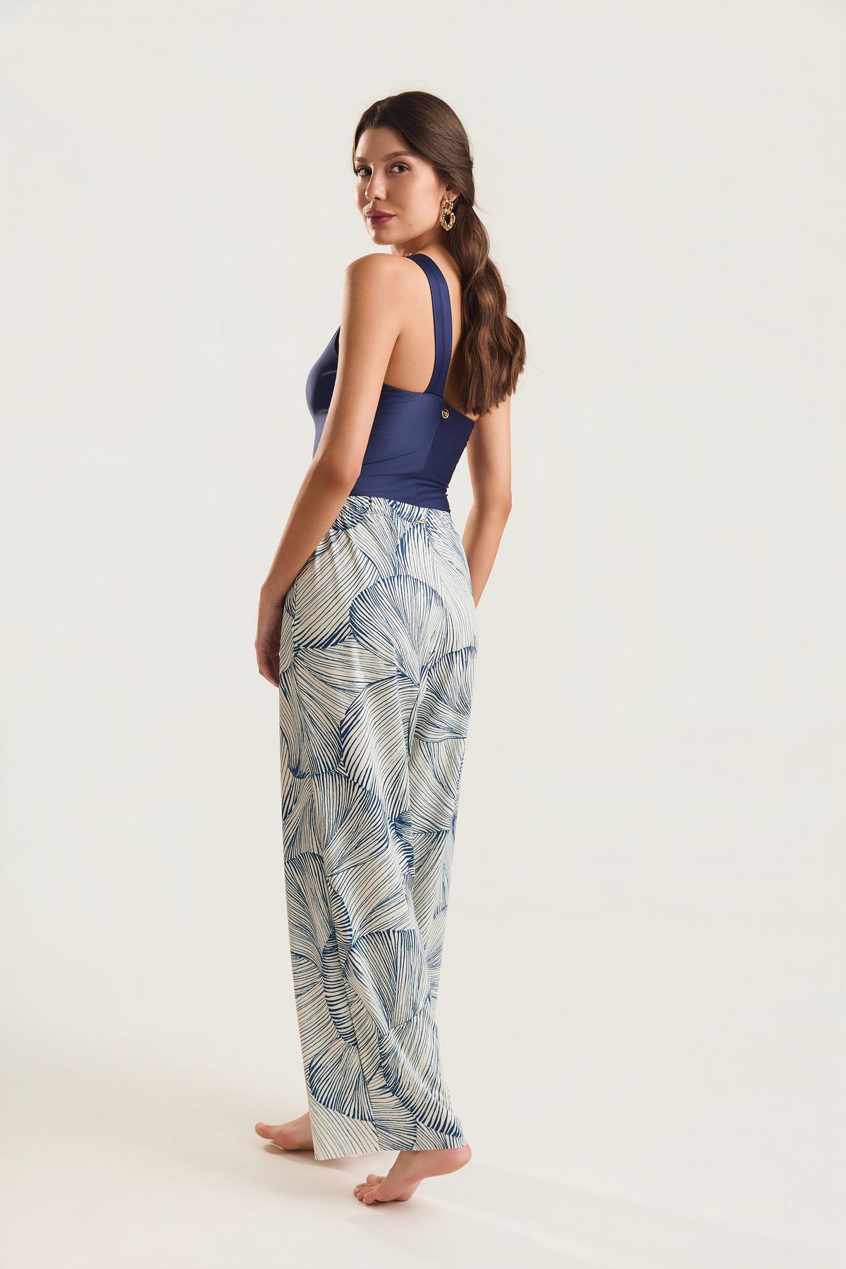 Calça pantalona estampada com conchas azuis, modelo de costas em postura de três quartos com leve torção do tronco e olhar sobre o ombro